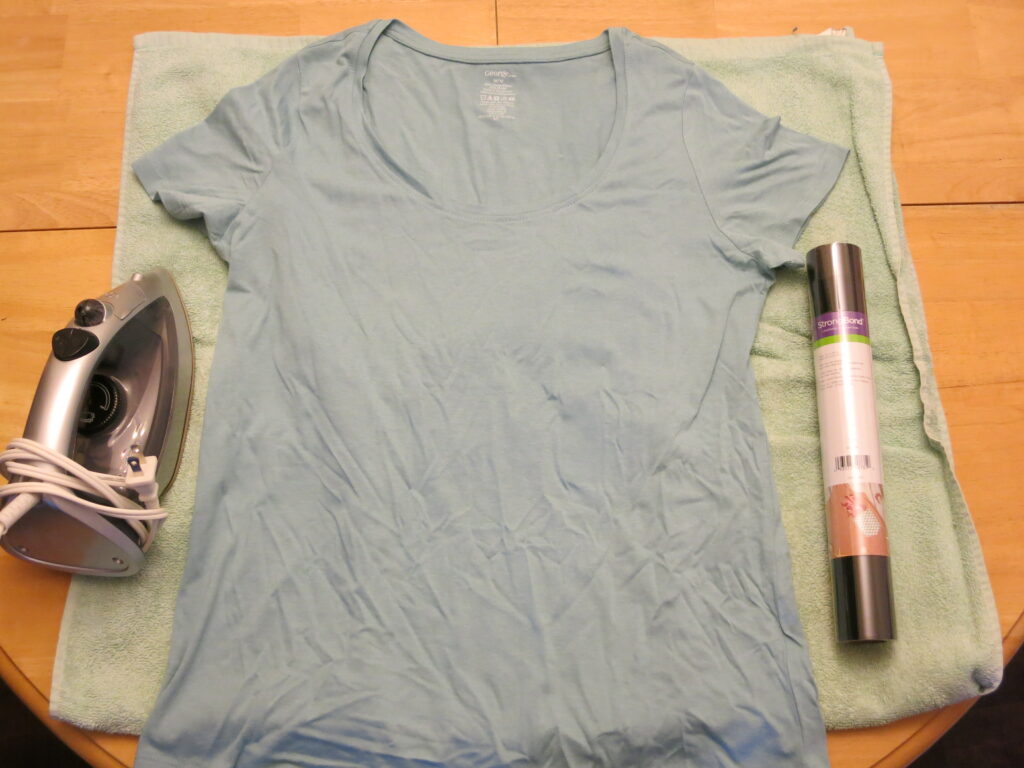 An iron, a blue T-shirt, and a roll of black iron-on vinyl; with a green towel underneath. 