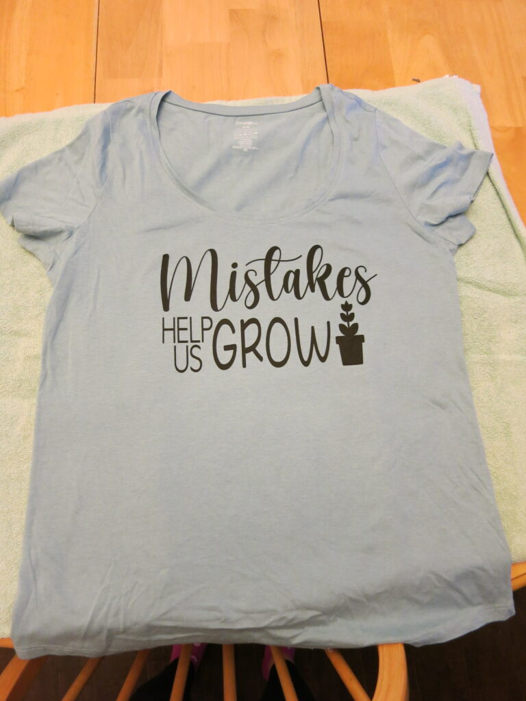 A blue T-shirt on top of a green towel. The T-shirt has black writing that says "Mistakes help us grow" with a picture of a black potted plant beside it. 