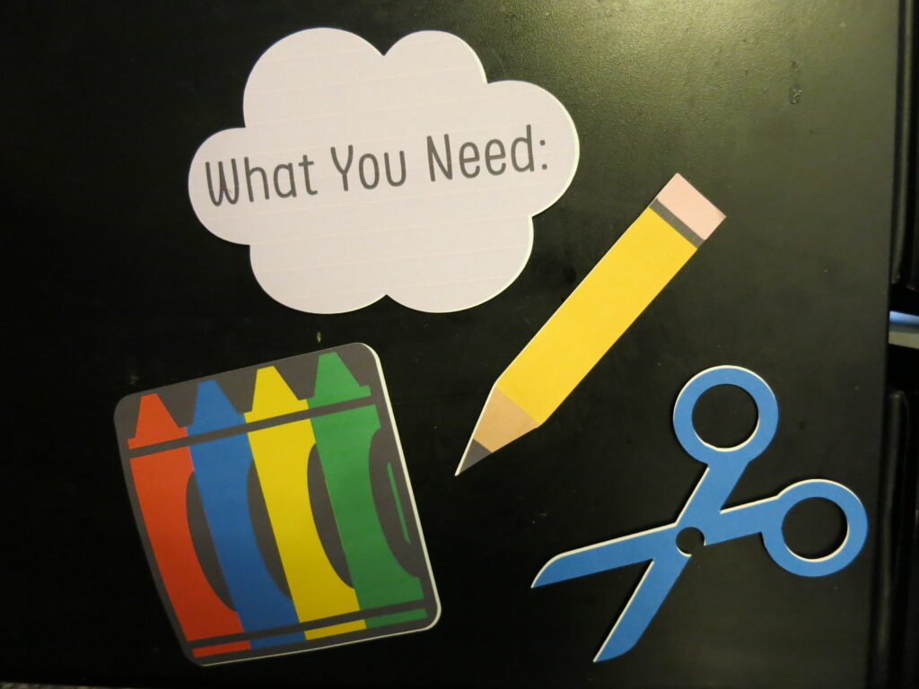 Four magnets on a black fridge. The magnets are crayons, a pencil, scissors, and a purple cloud that says "What You Need". 