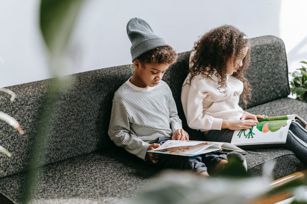 Two children, a boy and a girl, sitting on a couch together each reading a picture book. 