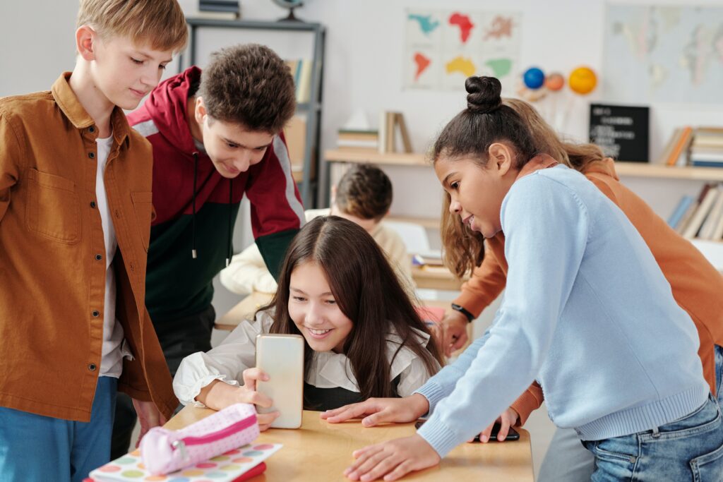 One female stident looking a smartphone while four other students surround her to look as well. They are all in a classroom. 